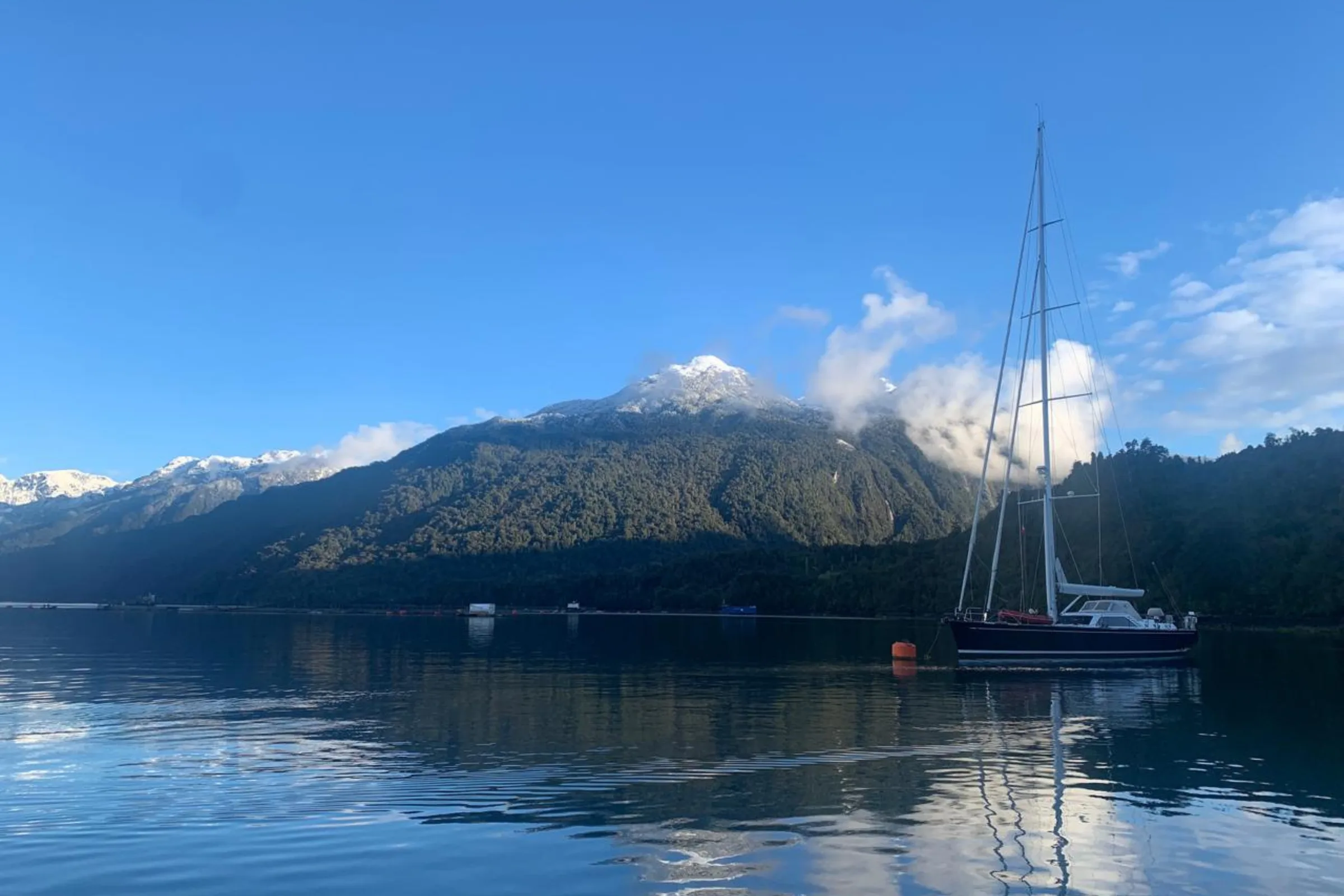Velero fondeado en un fiordo patagónico, con montaña boscosa y cumbre nevada de fondo
